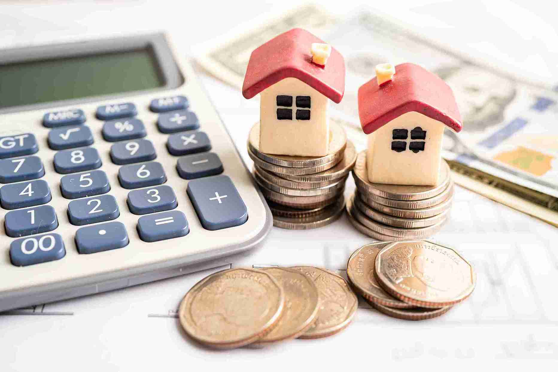 Links liegt ein Taschenrechner, rechts daneben ein Haufen Geld und Münzen mit kleinen Häusern