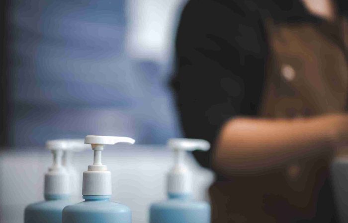 Shampoo nach Haartransplantation – wichtige Pflegehinweise 1 Mehrere blaue Spenderflaschen mit weißem Pumpverschluss, stehen für Shampoo nach Haartransplantation, in unscharfem Hintergrund einer Person in einem Salon oder einer Klinik.
