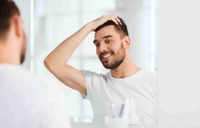 Pflege nach Haartransplantation: So gelingt die Heilung 1 Ein lächelnder Mann betrachtet sein Haar im Spiegel und überprüft das Ergebnis seiner Behandlung – Symbolbild für die richtige Pflege nach Haartransplantation.