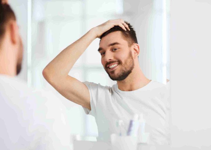 Pflege nach Haartransplantation: So gelingt die Heilung 1 Ein lächelnder Mann betrachtet sein Haar im Spiegel und überprüft das Ergebnis seiner Behandlung – Symbolbild für die richtige Pflege nach Haartransplantation.