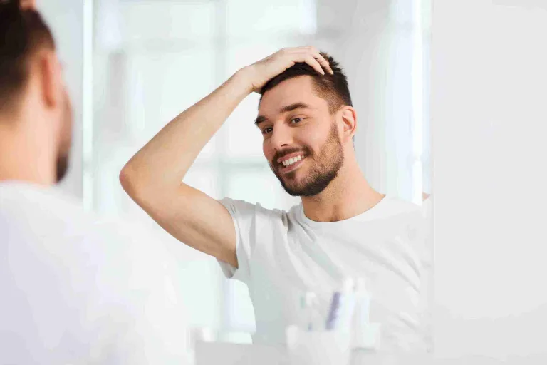 Ein lächelnder Mann betrachtet sein Haar im Spiegel und überprüft das Ergebnis seiner Behandlung – Symbolbild für die richtige Pflege nach Haartransplantation.