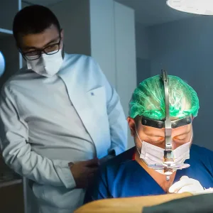 Arzt im weißen Kittel beaufsichtigt eine Fachkraft mit Stirnlampe während der Behandlung