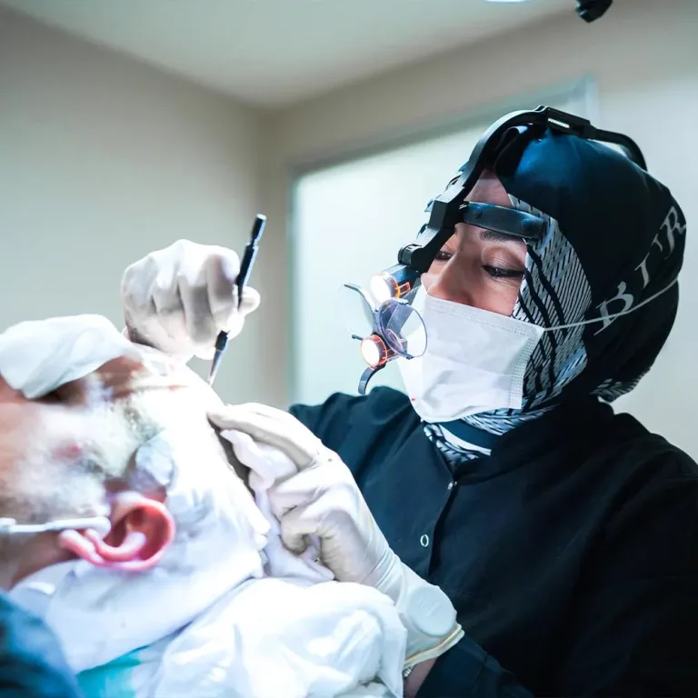 Chirurgin mit Kopftuch und Lupenbrille hält ein Instrument an der Stirn eines liegenden Patienten